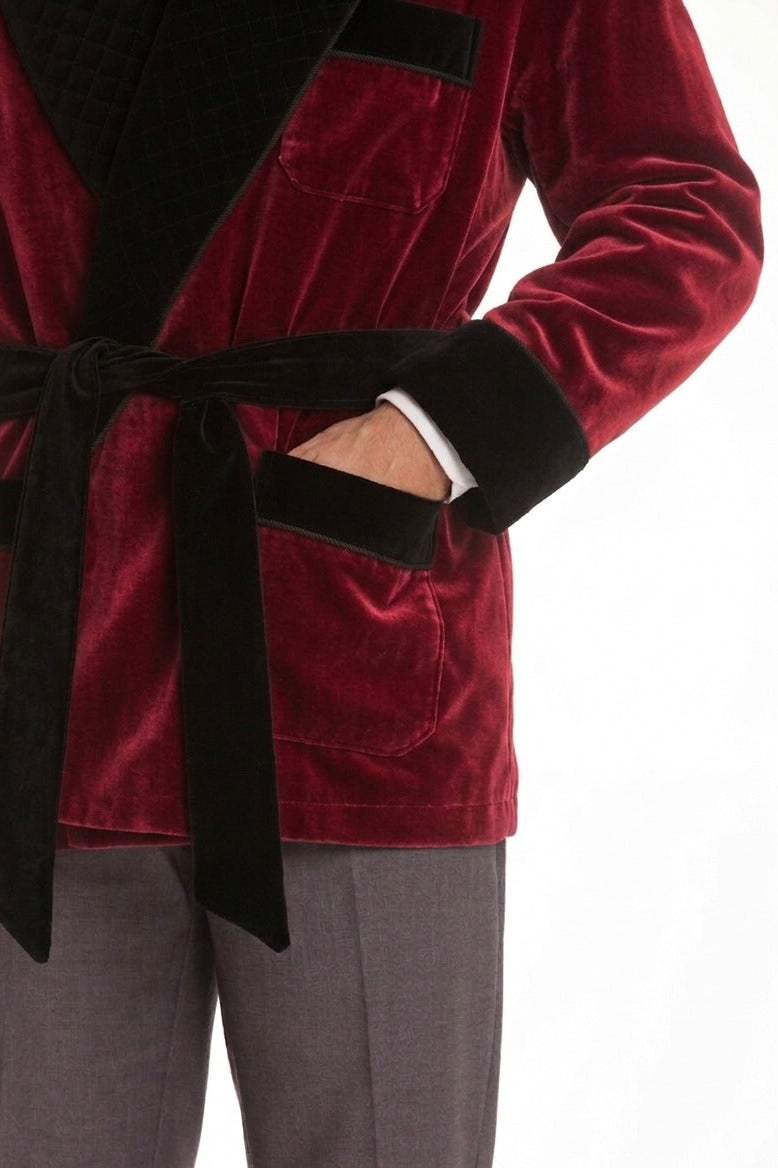 A close-up of a person wearing a plush red velvet robe with black trim and a matching black tie belt, paired with gray trousers, against a plain white background.